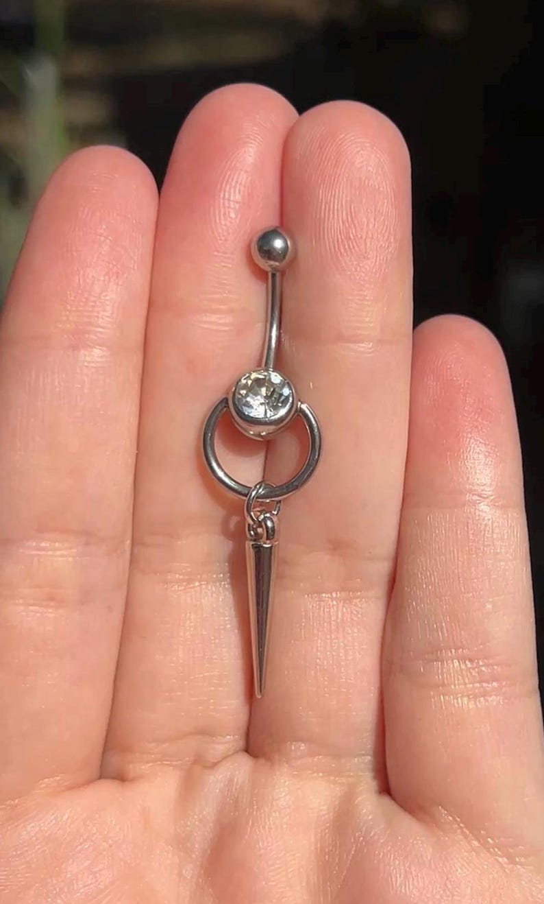 Peut inclure: Un piercing de nombril argent&eacute; compos&eacute; d'une barre courbe avec une petite boule sup&eacute;rieure et une grosse pierre ronde transparente &agrave; la base. Un anneau m&eacute;tallique est fix&eacute; &agrave; la pierre, suspendant une longue breloque en forme de c&ocirc;ne pointu.
