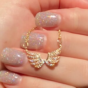 Op de afbeelding: Een goudkleurige hangerketting met een paar engelen vleugels, bezet met kleine, sprankelende kristallen. De vleugels zijn verbonden door een fijne ketting. De achtergrond is wazig, waardoor de sieraden worden benadrukt.