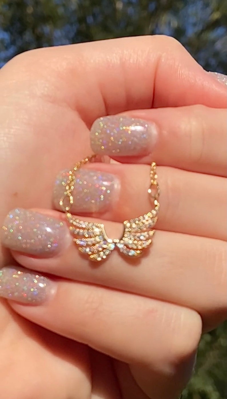 Op de afbeelding: Een goudkleurige hangerketting met een paar engelen vleugels, bezet met kleine, sprankelende kristallen. De vleugels zijn verbonden door een fijne ketting. De achtergrond is wazig, waardoor de sieraden worden benadrukt.