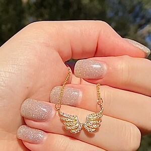 Op de afbeelding: Een goudkleurige ketting met een hanger in de vorm van engelen vleugels, bezet met kleine, sprankelende kristallen. De ketting wordt tegen een vage groene achtergrond gehouden, wat het delicate ontwerp en de ingewikkelde details van de sieraden benadrukt.