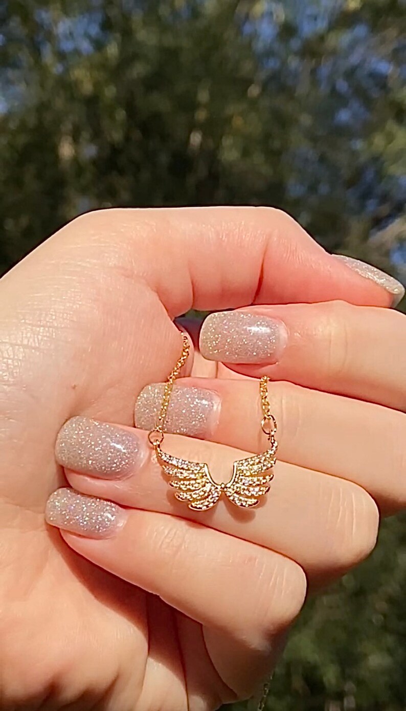 Op de afbeelding: Een goudkleurige ketting met een hanger in de vorm van engelen vleugels, bezet met kleine, sprankelende kristallen. De ketting wordt tegen een vage groene achtergrond gehouden, wat het delicate ontwerp en de ingewikkelde details van de sieraden benadrukt.