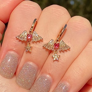 May include: Gold-coloured hoop earrings. Each earring has a wing design with clear and pink gemstones, a heart-shaped pink gem, and a dangling star charm. The earrings are shown against a blurred background.