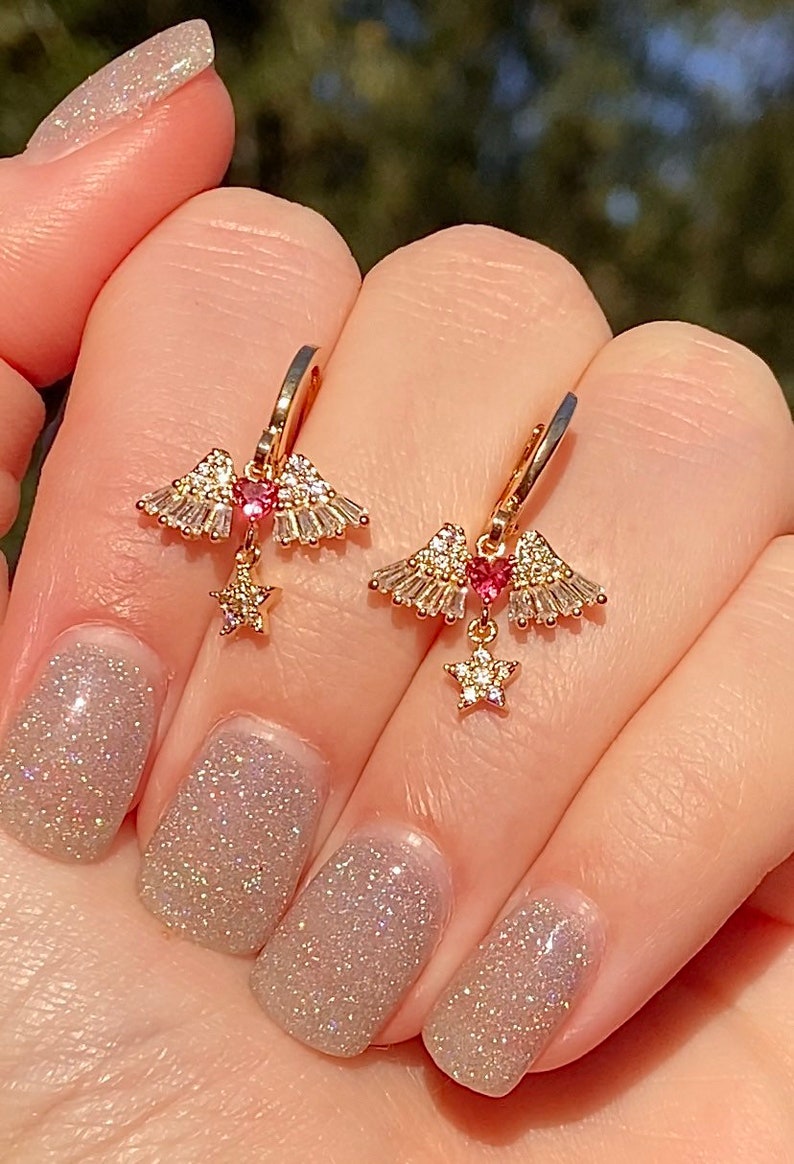 May include: Gold-coloured hoop earrings. Each earring has a wing design with clear and pink gemstones, a heart-shaped pink gem, and a dangling star charm. The earrings are shown against a blurred background.