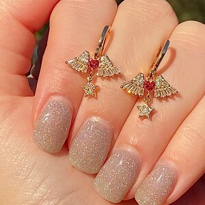 May include: Gold-coloured earrings with a distinctive design. Each earring has a cluster of small, sparkling crystals, a small red gem, and a dangling star-shaped charm. The earrings are held up by a hand with glittery nail polish.