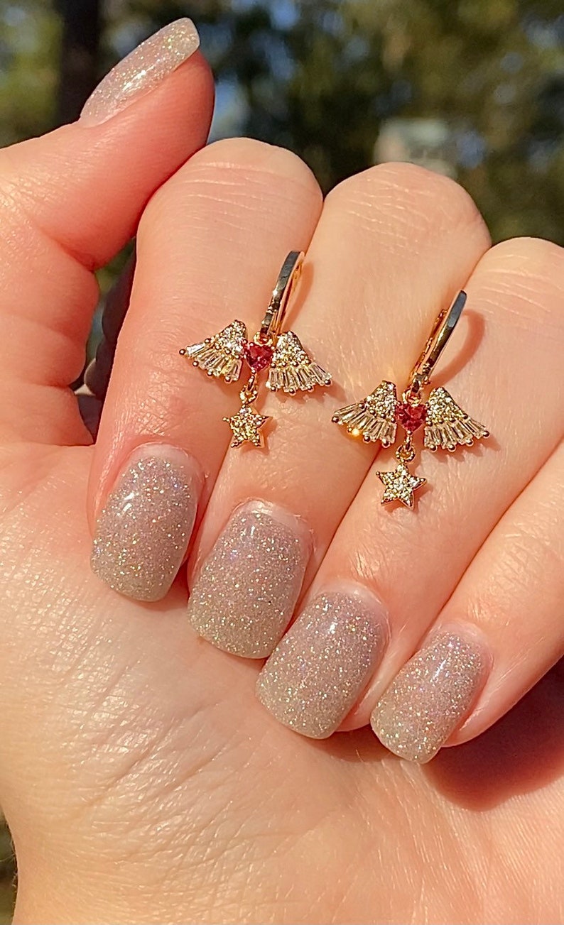 May include: Gold-coloured earrings with a distinctive design. Each earring has a cluster of small, sparkling crystals, a small red gem, and a dangling star-shaped charm. The earrings are held up by a hand with glittery nail polish.