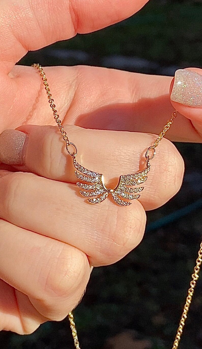 Op de afbeelding: Een goudkleurige ketting met een paar engelen vleugels, elke vleugel versierd met kleine, sprankelende kristallen. De ketting wordt door een persoon vastgehouden, waarbij de fijne ketting en het gedetailleerde ontwerp van de hanger worden benadrukt. De vleugels zijn ongeveer 2 cm breed.