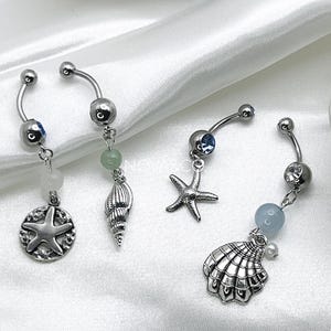 May include: Three silver belly button rings with different charms. The charms are a seashell, a starfish, and a seashell with a pearl. The rings are on a white background.