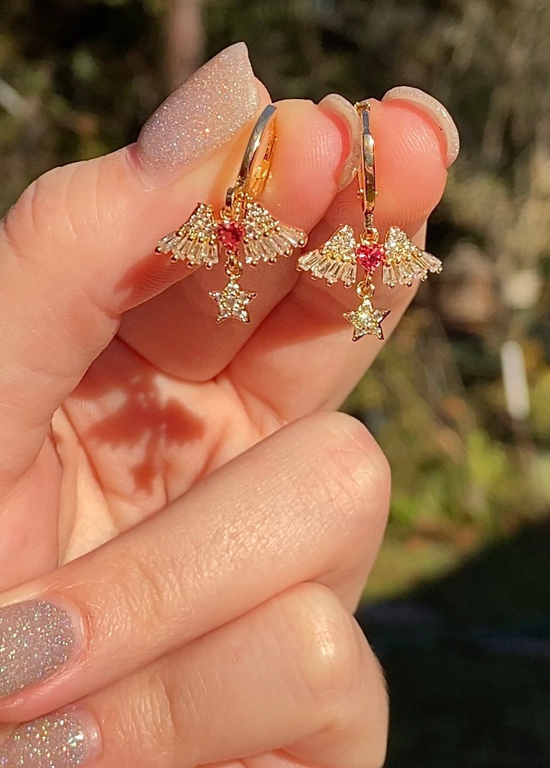 May include: Gold hoop earrings with a celestial design. Each earring has a central red gemstone, surrounded by a fan of clear, baguette-cut stones. A dangling star charm with clear stones hangs below.
