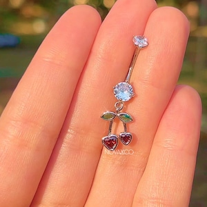 May include: A silver belly button ring with a cherry design. The ring features a clear gem at the top, a larger blue gem in the middle, and two red heart-shaped gems at the bottom, with green leaves. The brand name "NOVACXO" is visible.