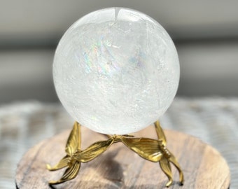 May include: A white crystal ball with iridescent shimmer sits on a gold metal stand with a butterfly design. The stand is on a wooden base.