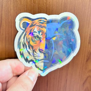 May include: A die-cut sticker featuring a split-face design of a tiger. The left side displays a realistic orange and black tiger face, while the right side shows a dark, holographic geometric silhouette of a bear with a glowing yellow eye.
