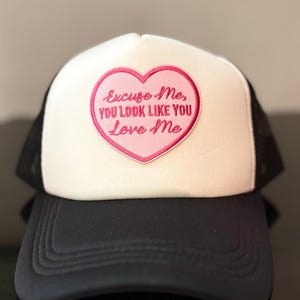 Puede incluir: Gorra de camionero en blanco y negro con un parche rosa en forma de corazón. El parche presenta la frase bordada: "Excuse Me, You Look Like You Love Me". La gorra tiene una visera curva y una parte trasera de malla.