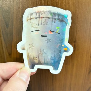 May include: A die-cut sticker featuring a stylized, anthropomorphic pillow character with a blue and grey gradient finish. The pillow has a stitched top edge, closed eyes, a small mouth, and star patterns. The text SWEET DREAMS is printed on the left.