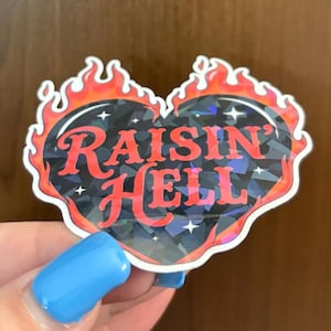 May include: A heart-shaped sticker with the words "Raisin' Hell" in red script. The heart has a black, textured center with a holographic effect and is outlined with flames.