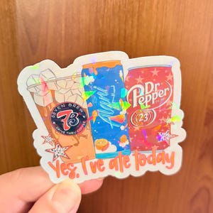 May include: A holographic sticker featuring three beverage containers: a Seven Brew iced coffee cup, a blue Alani Nu energy drink can, and a red Dr. Pepper can. The sticker includes orange text at the bottom that reads, Yes, I've ale today.