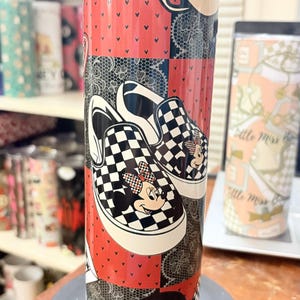 May include: A tall, insulated tumbler featuring a patchwork design with red, black, and white elements. The design includes a pair of black and white checkered sneakers with Minnie Mouse graphics. The tumbler is cylindrical and stands on a black base.