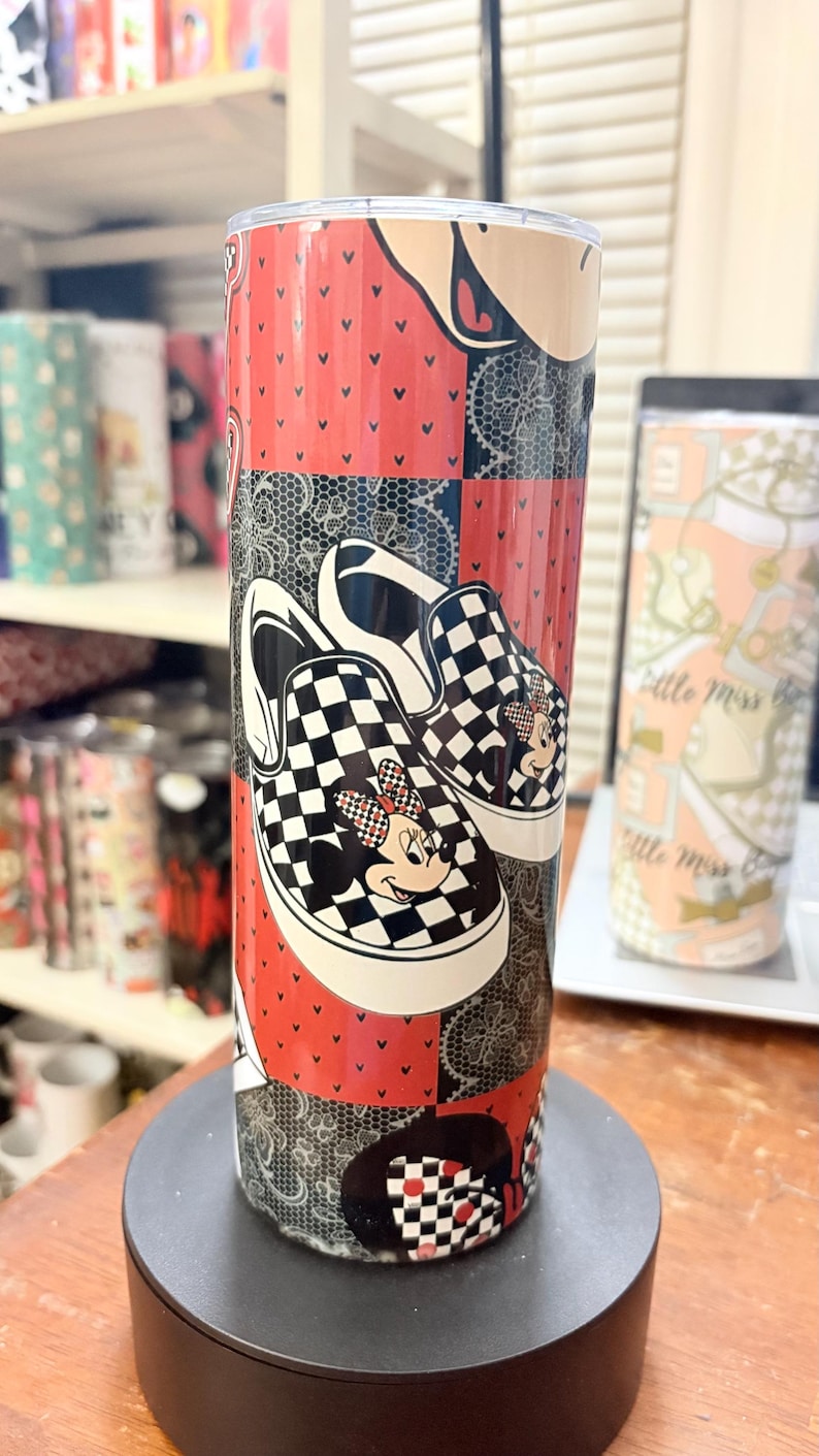 May include: A tall, insulated tumbler featuring a patchwork design with red, black, and white elements. The design includes a pair of black and white checkered sneakers with Minnie Mouse graphics. The tumbler is cylindrical and stands on a black base.