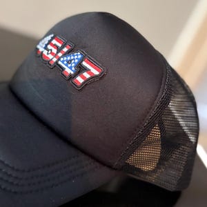 May include: Black trucker hat with a mesh back and a curved brim. The hat features a patch with the number "4547" in red, white, and blue, resembling the American flag. The hat is made of a durable material.