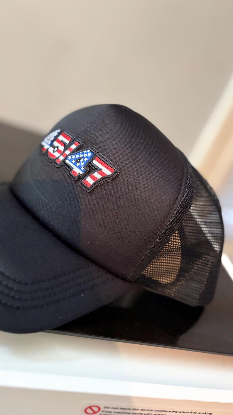May include: Black trucker hat with a mesh back and a curved brim. The hat features a patch with the number "4547" in red, white, and blue, resembling the American flag. The hat is made of a durable material.