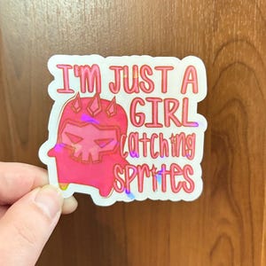 May include: A die-cut sticker with a holographic finish featuring a pink, stylized, horned character and the text "I'M JUST A GIRL CATCHING SPRITES" in pink and red lettering. The design has a white border and a reflective, iridescent surface.