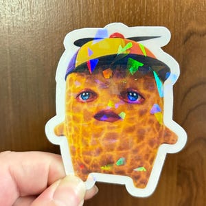 May include: A die-cut sticker featuring a textured, golden-brown peanut character with large blue eyes and a small mouth. The character wears a blue and yellow propeller cap. The sticker has a holographic finish with scattered geometric light reflections.