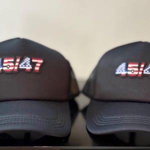 May include: Two black trucker hats with the text "45/47" in red, white, and blue. The hats have a mesh back and a curved brim. The hats are displayed on a black surface.