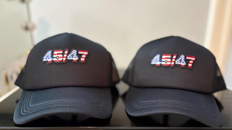 May include: Two black trucker hats with the text "45/47" in red, white, and blue. The hats have a mesh back and a curved brim. The hats are displayed on a black surface.