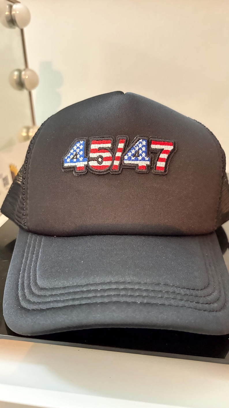 May include: Black trucker hat with the number "45/47" embroidered on the front in red, white, and blue. The hat has a mesh back and a curved brim. A casual accessory.