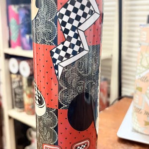 May include: A tall, stainless steel tumbler with a patchwork design. The tumbler features red, black, and white patterns, including a checkered design, lace, and a logo. The tumbler is cylindrical and has a clear lid.
