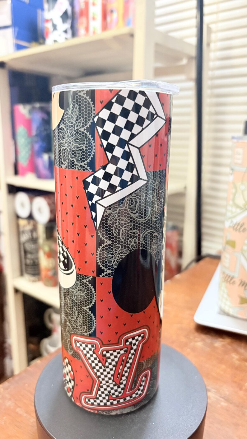 May include: A tall, stainless steel tumbler with a patchwork design. The tumbler features red, black, and white patterns, including a checkered design, lace, and a logo. The tumbler is cylindrical and has a clear lid.