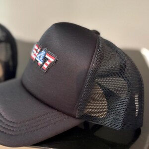 May include: Black trucker hat with a mesh back and a curved brim. The front of the hat features a patch with the number "47" in red, white, and blue, resembling the American flag. The hat is made of a durable fabric.