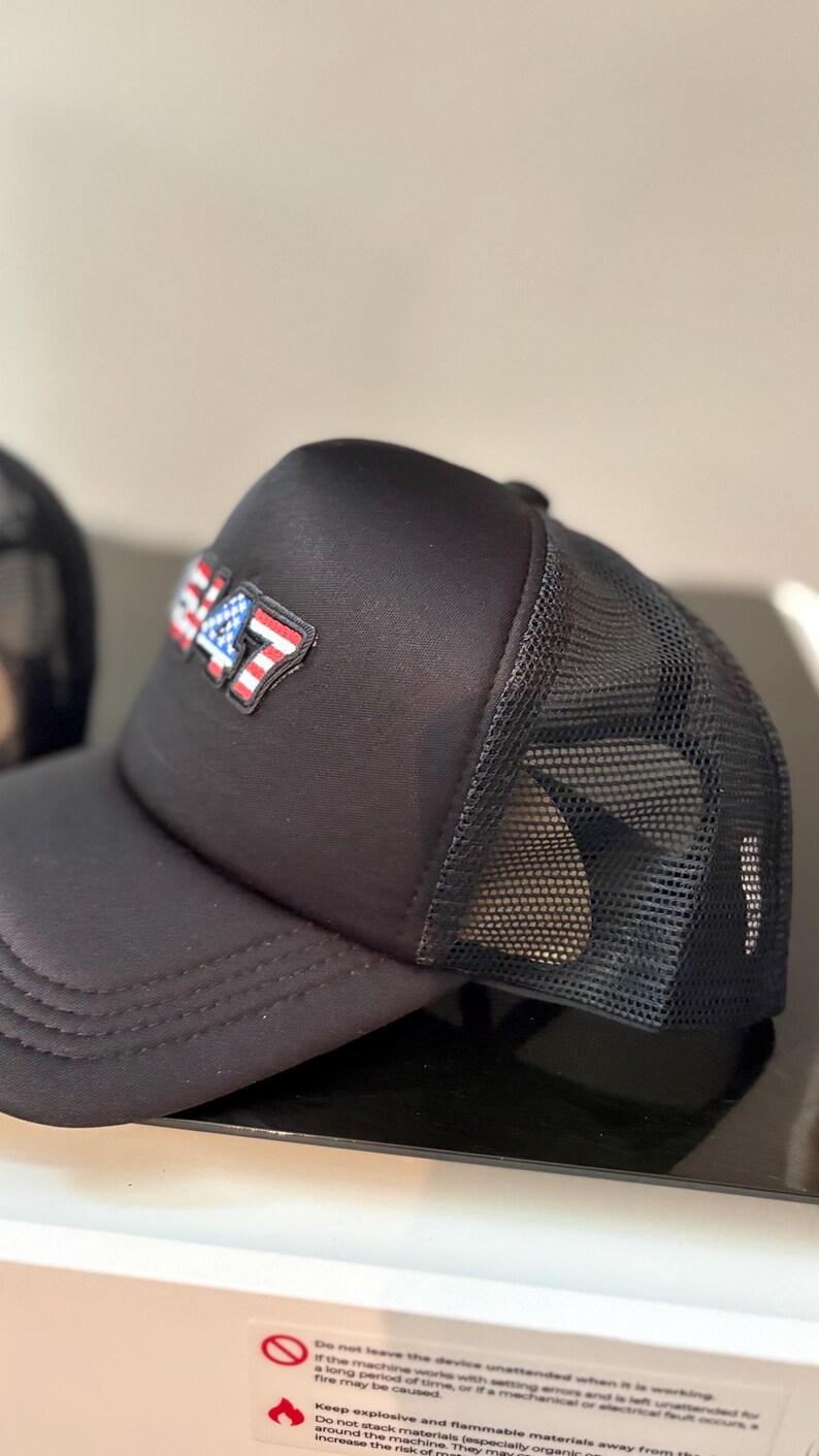 May include: Black trucker hat with a mesh back and a curved brim. The front of the hat features a patch with the number "47" in red, white, and blue, resembling the American flag. The hat is made of a durable fabric.