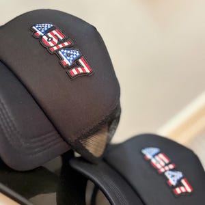 May include: Black trucker hats with the text "45/47" in a red, white, and blue design, resembling the American flag. The hats have a mesh back and a curved brim. The hats are displayed on a reflective surface.