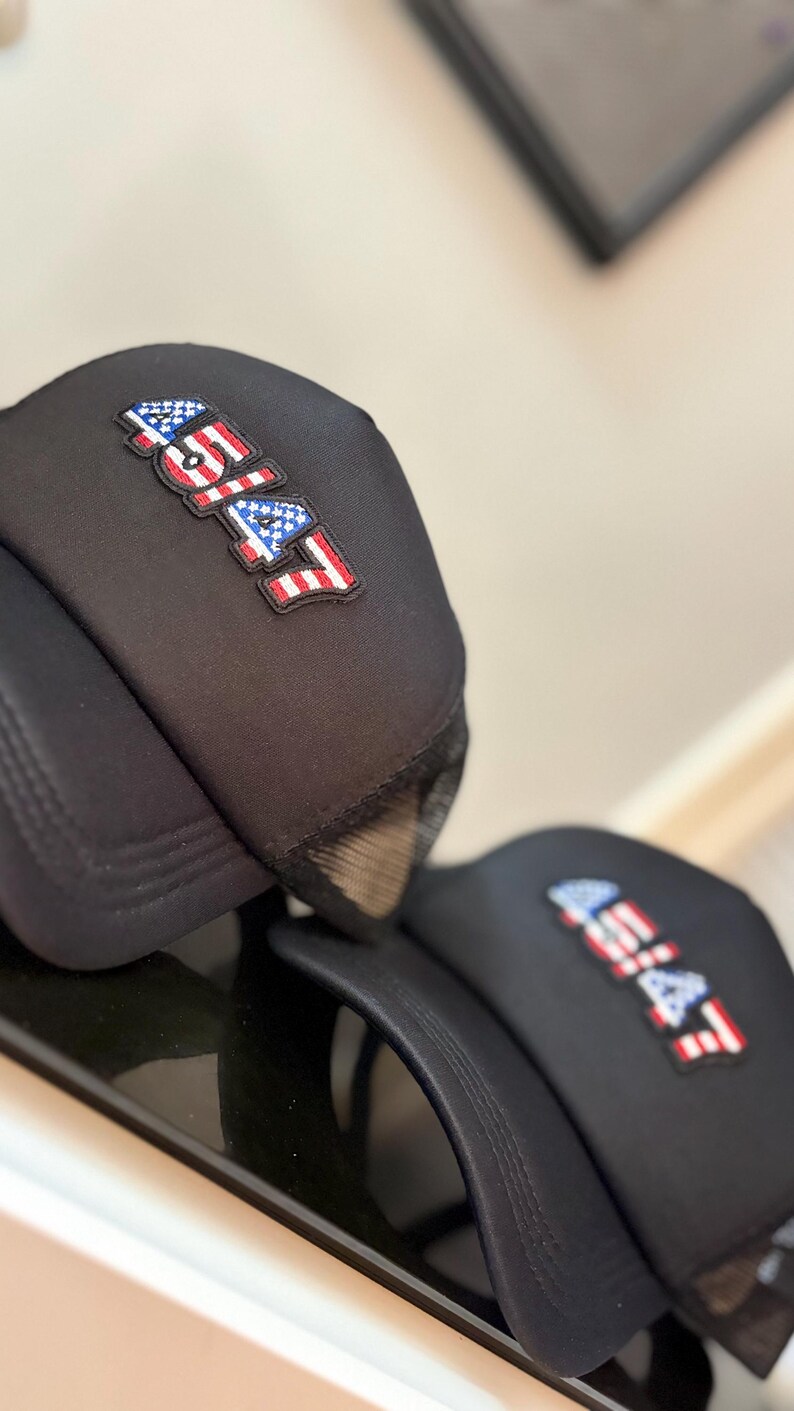 May include: Black trucker hats with the text "45/47" in a red, white, and blue design, resembling the American flag. The hats have a mesh back and a curved brim. The hats are displayed on a reflective surface.
