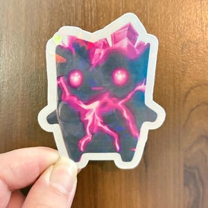 May include: A die-cut sticker featuring a dark, blocky creature with glowing pink eyes and bright pink lightning-like patterns across its body. The top of the figure is adorned with crystalline shapes, and the entire design has a holographic finish.
