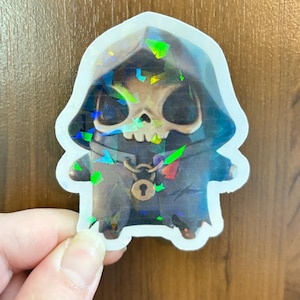May include: A holographic sticker featuring a stylized, small, hooded figure with a skull face. The character wears a dark cloak with a prominent circular lock pendant hanging from a chain around its neck. The surface has a reflective finish.