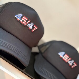 May include: Two black trucker hats with a mesh back and a curved brim. Each hat features a patch with the number "45/47" in red, white, and blue, resembling the American flag. The hats are displayed on a white surface.