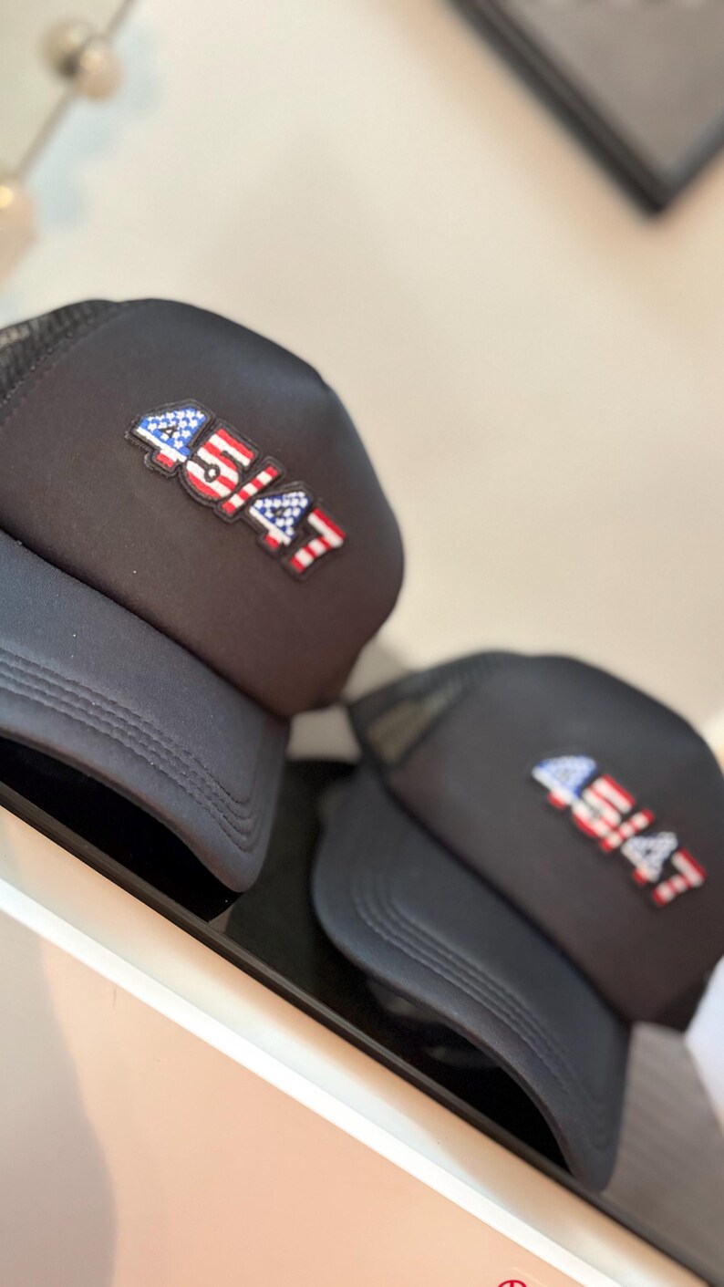 May include: Two black trucker hats with a mesh back and a curved brim. Each hat features a patch with the number "45/47" in red, white, and blue, resembling the American flag. The hats are displayed on a white surface.