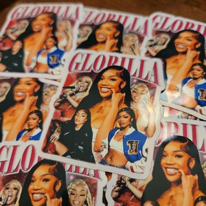 May include: A collection of stickers featuring a smiling woman with long dark hair. The stickers have the word "GLORILLA" in a stylized font. The background of the stickers includes images of the woman in various poses, wearing a blue and white jacket.