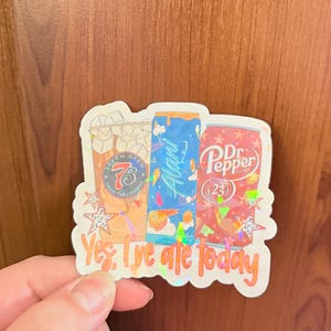 May include: A holographic sticker featuring three beverage containers: a Seven Brew iced coffee, an Alani Nu energy drink, and a Dr. Pepper can. The sticker is decorated with star shapes and includes the text "Yes, I've ate today" in orange lettering.
