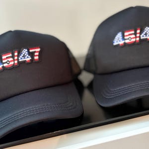 May include: Two black trucker hats with the text "45/47" in red, white, and blue, resembling the American flag. The hats have a curved brim and a mesh back.