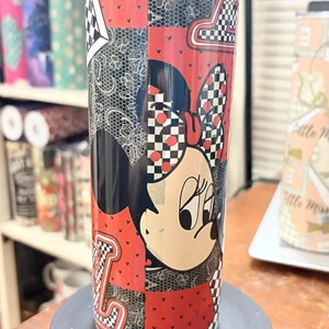 May include: A stainless steel tumbler featuring a patchwork design with Minnie Mouse. The tumbler has a red, black, and white color scheme with a checkered pattern and the letter "L".