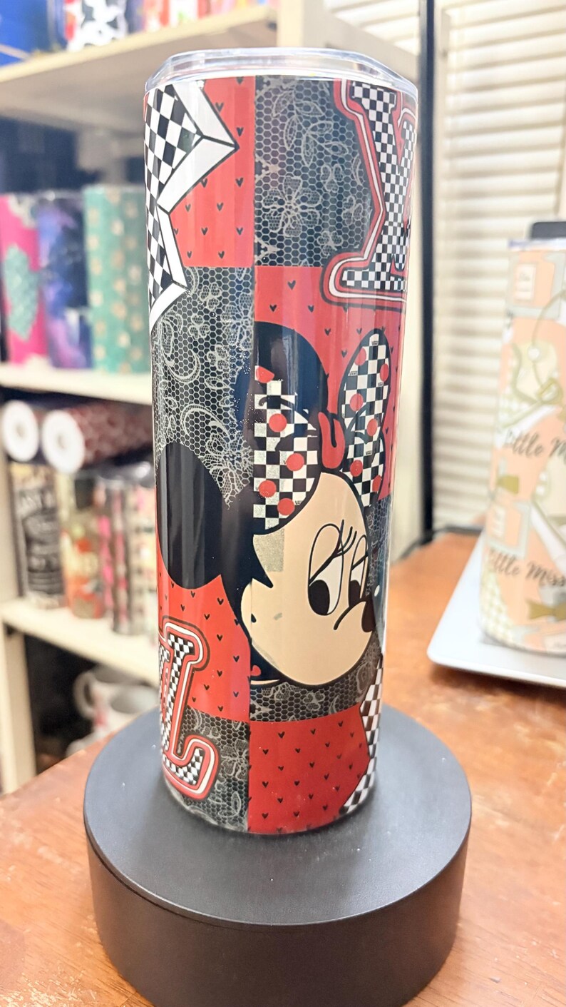 May include: A stainless steel tumbler featuring a patchwork design with Minnie Mouse. The tumbler has a red, black, and white color scheme with a checkered pattern and the letter "L".