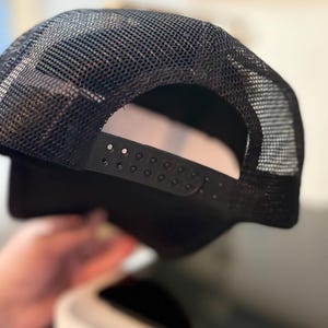 May include: Black trucker hat with a mesh back and adjustable snap closure. The hat features a solid black front panel and a curved brim. The mesh back allows for breathability, making it suitable for various activities.