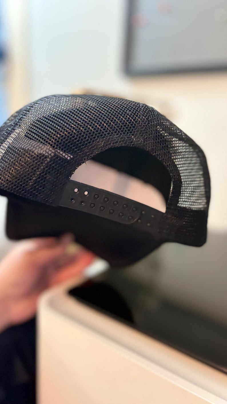 May include: Black trucker hat with a mesh back and adjustable snap closure. The hat features a solid black front panel and a curved brim. The mesh back allows for breathability, making it suitable for various activities.