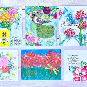 May include: Eight colorful cards with floral designs and inspirational quotes. Each card features a different flower, including sunflowers, lilies, and roses. The quotes are handwritten in black ink and include verses from the Bible.