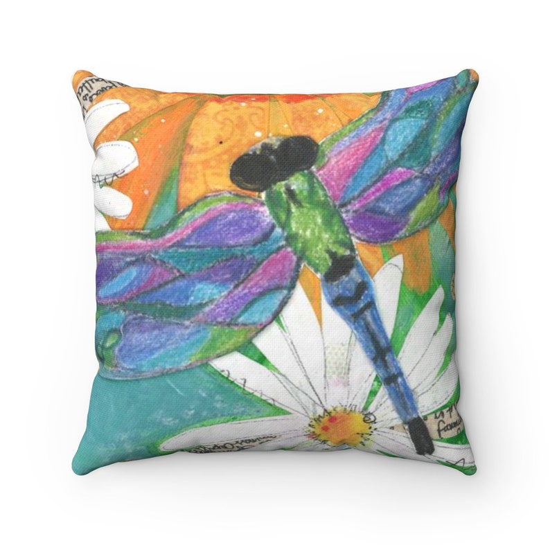 May include: A colorful pillow featuring a dragonfly with blue, green, purple, and pink wings. The dragonfly is perched on a white daisy with a yellow center. The background is a mix of orange, yellow, and green.