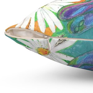 May include: A decorative pillow with a floral design featuring white daisies with yellow centers, green leaves, and purple accents. The pillow has a zipper closure and a white background.