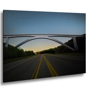 May include: A photograph of a bridge over a road at night. The bridge is a light gray color and has an arched design. The road has yellow lines down the center. The sky is dark blue and filled with stars.