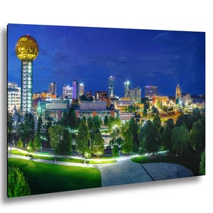 Puede incluir: Paisaje urbano nocturno con una torre esférica dorada y edificios iluminados. Zonas verdes y árboles en primer plano, bajo un cielo azul oscuro. La imagen muestra una vista panorámica de una ciudad.